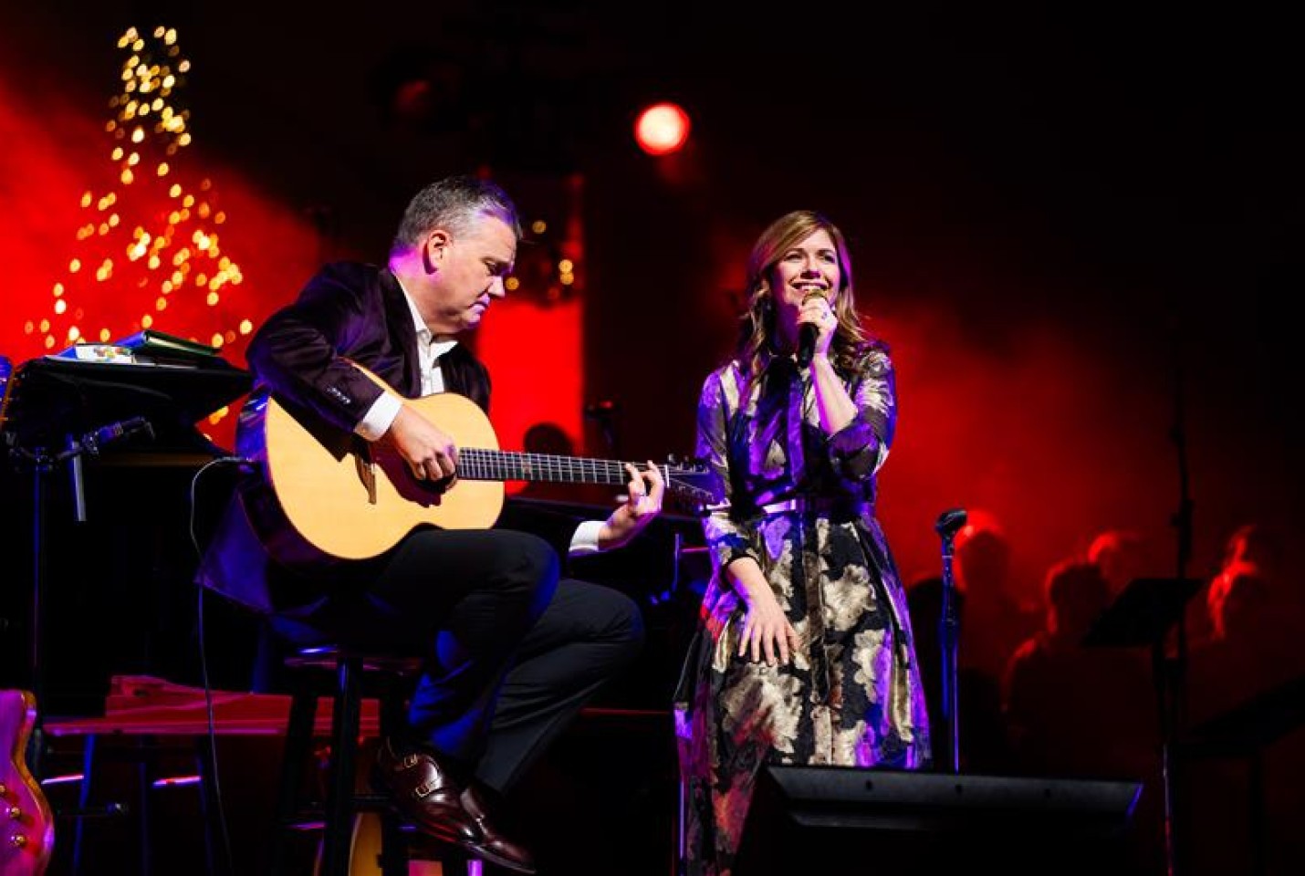 Keith and Kristyn Getty sitting down on chairs on a Stage performing. Keith holds an acoustic guitar and Kristyn is holding a Microphone.